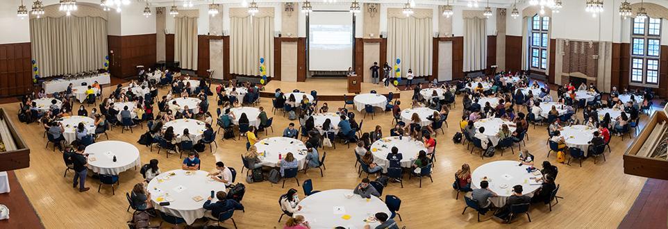 Panoramic view of a U-M campus event in a ballroom, with attendees seated at round tables and a speaker presenting at the front.