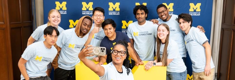 Group of students in "Transfer Connections" shirts posing for a selfie in front of a University of Michigan backdrop.